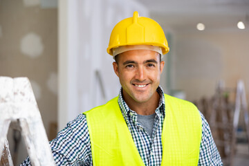 Portrait of positive man builder in vest and helmet standing in apartment during repair works, looking at camera and smiling.