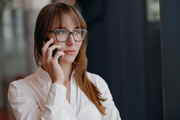 A focused and determined young woman wearing glasses is talking on her phone in a modern and stylish setting