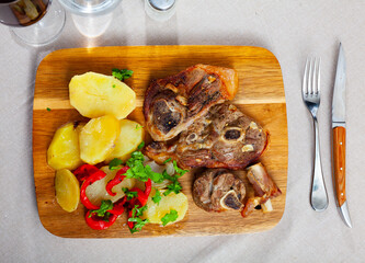 Appetizing pieces of roasted lamb leg with side dish of baked potatoes and red pepper served on wooden board with fresh greens..