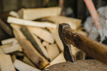 A sharp axe is embedded in a sturdy log. In the background, there are stacked firewood pieces. It...