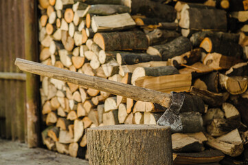 A sharp axe is embedded in a sturdy log. In the background, there are stacked firewood pieces. It...