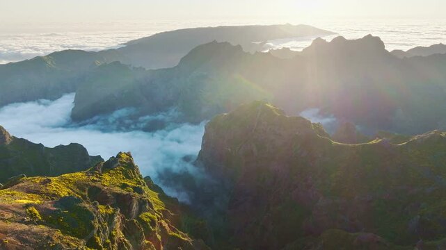 Aerial view at Pico do Arieiro of mountains over clouds with blooming Cytisus shrubs on sunset with sunburst. Madeira island, Portugal. Pull away out dolly retreat shot