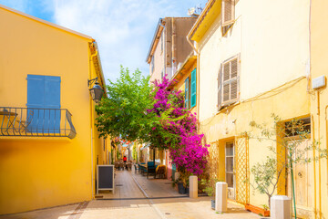 A picturesque alley with purple Bougainvillea flowers covering the entry and a sidewalk cafe and shops in the old town village section of Saint-Tropez, France, on the French Riviera.	