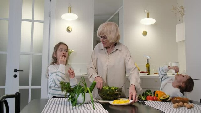 Grandma and her grandchildren cook in a bright modern kitchen. The grandmother passes one of the young girls a plate with chopped bell peppers, while the other girl is already enjoying a piece of