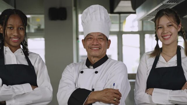 Portrait of happy professional Asian senior male chef teacher in uniform with chef hat and his multiracial students standing smiling and arms crossed while looking at camera in kitchen at restaurant. 