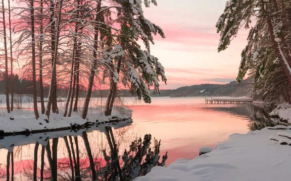 A serene winter landscape during sunset or sunrise. A calm body of water, possibly a lake or river, reflects the soft hues of the sky.