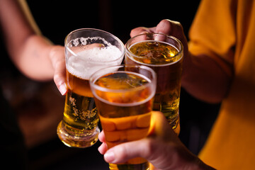 Close-up of friends clinking glasses of beer, celebrating together at a bar.The lively atmosphere captures laughter, camaraderie,good times, as young friends enjoy a night out with refreshing drinks