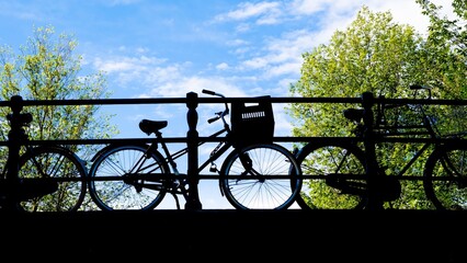 Bicycles parked on a bridge, in silhouette, with blue sky, light clouds and green tree leaves framing. Shot from low angle. Amsterdam, Holland, where residents bicycle as daily transportation.