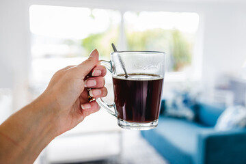 hand holding coffee in clear mug in front of blurred home interior background, habits and lifestyle concept
