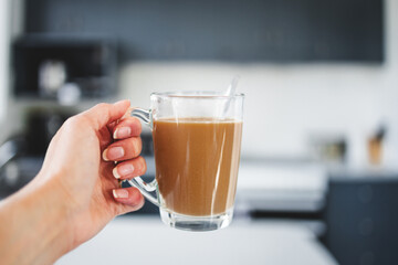 hand holding coffee in clear mug in front of blurred home interior background, habits and lifestyle concept
