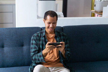 Obraz premium Young asian men wearing orange shirt and flannel is holding smart phone with smile on sofa.