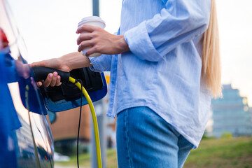Woman is connecting a charging cable to an electric car, enjoying a coffee while parked.