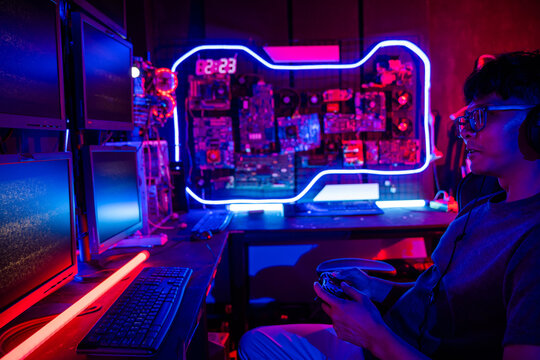 Young Asian man gamer wearing glasses and headphones playing a video game on modern gaming pc computer with purple light on night, plays online home games room on Hacker desktop dark background