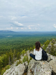 Naklejka premium A woman is comfortably sitting on a large rock while she enjoys an amazing view of a vast and beautiful forest stretching far and wide in the distance, surrounded by natures splendor