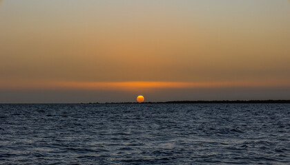 The sun, looking like an orange and yellow ball, setting and sinking into the horizon with orange lighting on the cloud bank above the horizon in Spencer gulf at Port Germein in South Australia.