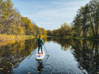 A man is riding a paddle board on a serene river, skillfully navigating the water while taking in the stunning natural scenery surrounding him, from lush trees to the open sky above