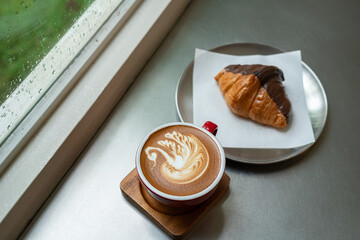 Hot coffee latte with latte art milk foam a swan shape in cup mug with chocolate Croissant on wood desk on top view.As breakfast In a coffee shop at the cafe,during business work concept,Rainy season