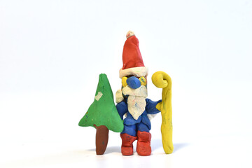 The figurine of Santa Claus with a bag of gifts on his back and a Christmas tree in his hands is made of plasticine on a white background.