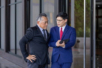 Asian senior businessman giving advice to the new manager and looking at information regarding sale record while working inside modern style office