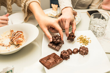 Two people sharing artisanal chocolates on a white plate, featuring decorative shapes and a variety of textures, with an accompanying dessert and drink in the background