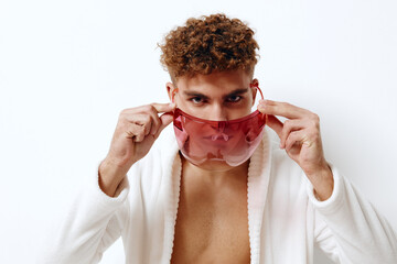 Stylish young man with curly hair wearing a red transparent face mask, posing confidently in a white robe against a clean, bright background. Fashion and self-expression concept.