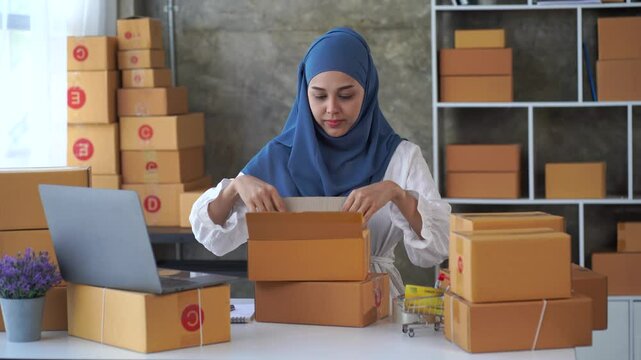 Entrepreneurial Woman Packing Order: A focused Muslim woman in a hijab carefully packs an order amidst a stack of shipping boxes, showcasing dedication and the dynamism of online business.  