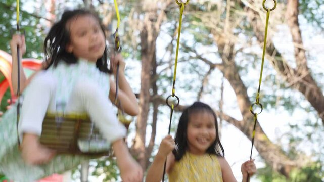 Cute sisters are having fun playing on the swings in a playground in a park. Children playing in the playground during summer vacation.