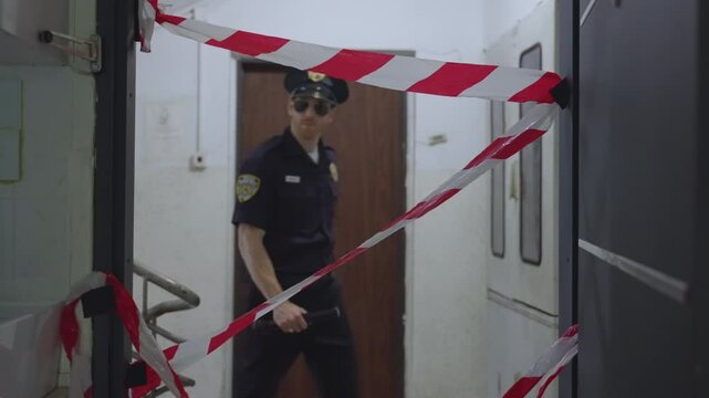 Policeman walking into apartment crime scene with caution police tape or ribbon. Officer or cop with blue uniform holding flashlight and wearing sunglasses enters room. Crossing blocked doorway