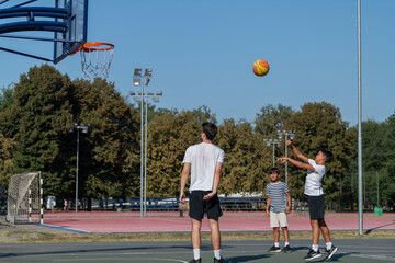 Boys engage in outdoor basketball training with a coach, practicing dribbling, passing, and shooting skills on a court surrounded by greenery under bright natural light, fostering teamwork