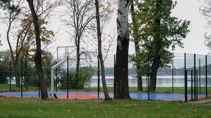 A deserted outdoor basketball court surrounded by autumn trees and a lake, highlighting solitude and seasonal change