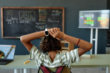 Back view of African American teenage girl stretching while sitting at desk in school classroom...