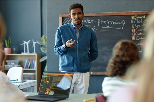 Waist up portrait of African American male teacher talking to group of children in school classroom and explaining IT programming copy space