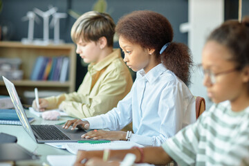 Fototapeta premium Side view of diverse group of children sitting in row in school classroom with focus on young African American girl using computer copy space