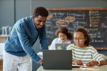 Fototapeta premium Portrait of friendly male teacher helping African American boy during lesson in school classroom and using laptop