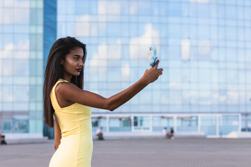 Fototapeta premium Indian woman in yellow dress taking a selfie with smartphone in front of a modern glass building