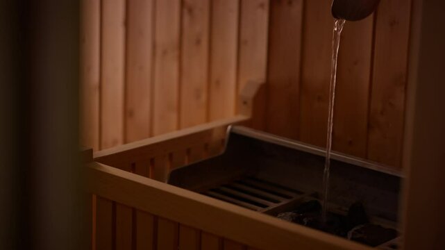 Close-up of water being poured onto hot sauna stones with steam rising, creating a relaxing atmosphere in a wooden sauna. Concept of wellness, rejuvenation, and traditional spa rituals.
