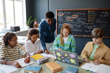 Wide angle view at young African American male teacher helping diverse group of children sitting at...