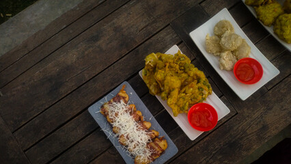 A top-down view of an assortment of Indonesian street food served on a rustic wooden table. A fried banana with grated cheese on top, a plate of vegetable fritters.
