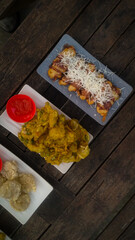 A top-down view of an assortment of Indonesian street food served on a rustic wooden table. A fried banana with grated cheese on top, a plate of vegetable fritters.