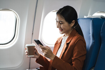 Smiling Asian woman enjoying her comfortable flight while sitting in the airplane cabin, wifi internet on the plane. Passengers near the window.