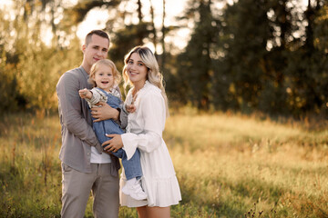Fototapeta premium Caucasian family of three, man, woman and child have fun walking in the park in summer. Family posing in the park at sunset with copy space.