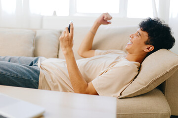 happy young man relaxing on the couch with smartphone, light colors, modern interior, cheerful expression, leisure time, cozy atmosphere, engaging with content, casual outfit, home comfort