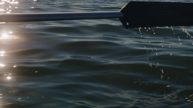 An oar splashing water with sunlight reflecting from the lake surface. Rowing a boat during sunset. 4K