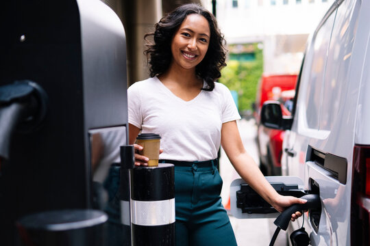Smiling woman holding coffee cup and charging electric car at station