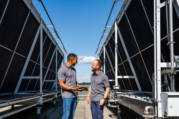 Two employees with digital tablet talking on rooftop beside refrigeration installation