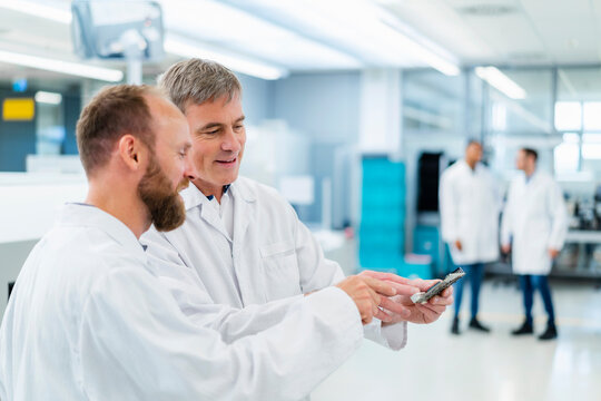 Technicians in electronics factory discussing over motherboard while colleagues are talking in background