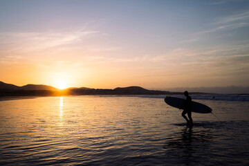 unrecognizable surfer silhouette with surfboard walking by the shore of the beach of Famara at sundown in Teguise, Lanzarote at Canary Islands in Spain, concept of wild nature and marine lifestyle