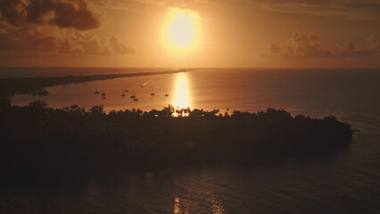 Golden sun illuminate calm tropical lagoon in French Polynesia, sunset sailboats anchored near a small island shore, serene and picturesque scene. Remote wild nature paradise, exotic summer travel