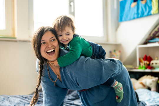 Cheerful mother giving piggyback ride to son on bed at home