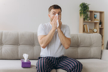 Unhappy middle aged man suffering from fever and flu, blowing nose in napkin, sitting in living...